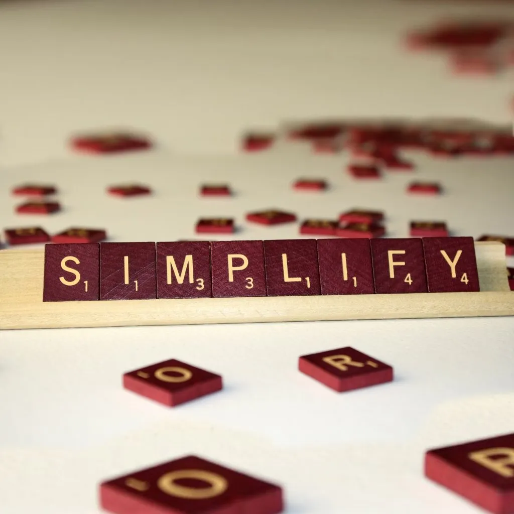 Maroon letter tiles spelling SIMPLIFY on a wooden tile rack, with loose tiles scattered around